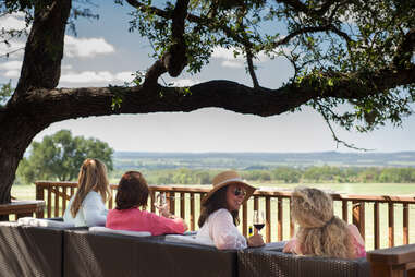 four women sit on couches overlooking a vineyard in texas wine country, under an oak tree