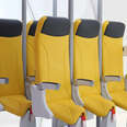 a prototype of a row of three standing airplane seats in a white mock airplane cabin. the seats are bright yellow and the bottoms resemble bicycle or horse saddles.